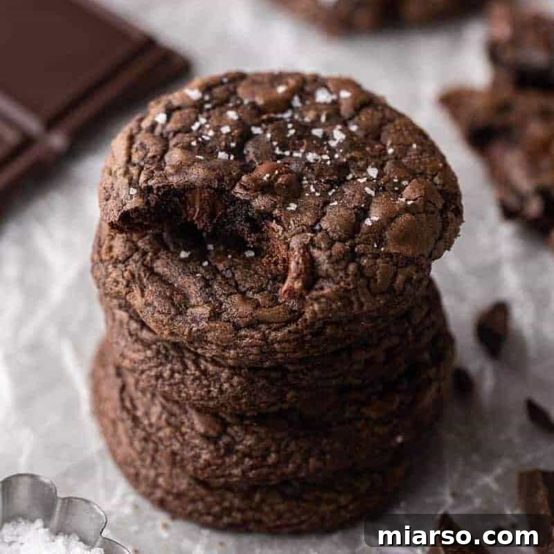 A delectable stack of four Crumbl brownie batter cookies, featuring perfect crinkly tops and delicate flaky sea salt, arranged beside gourmet dark chocolate squares and enticing chocolate shavings.