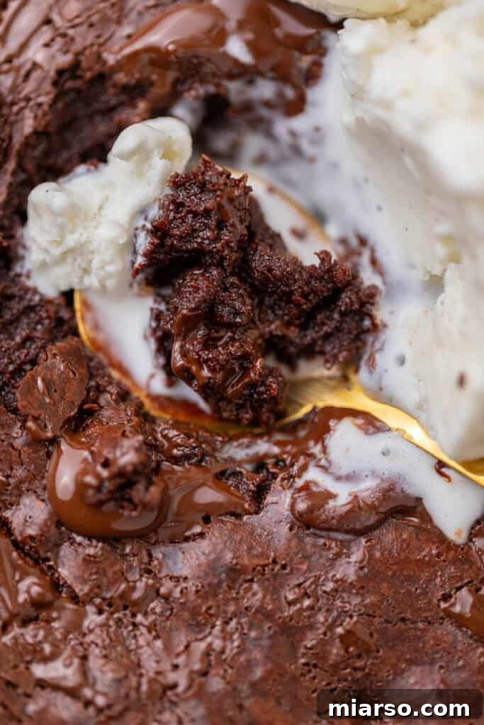Close-up of a warm, fudgy skillet brownie, gleaming with melted chocolate and adorned with scoops of vanilla ice cream gently melting on top, all being savored with a golden spoon.
