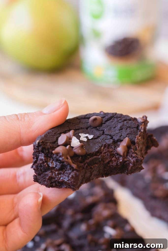 A hand holds a delectable vegan black bean brownie, generously topped with chocolate chips and flaky sea salt, revealing its dense, fudgy interior, with a fresh avocado subtly blurred in the background.