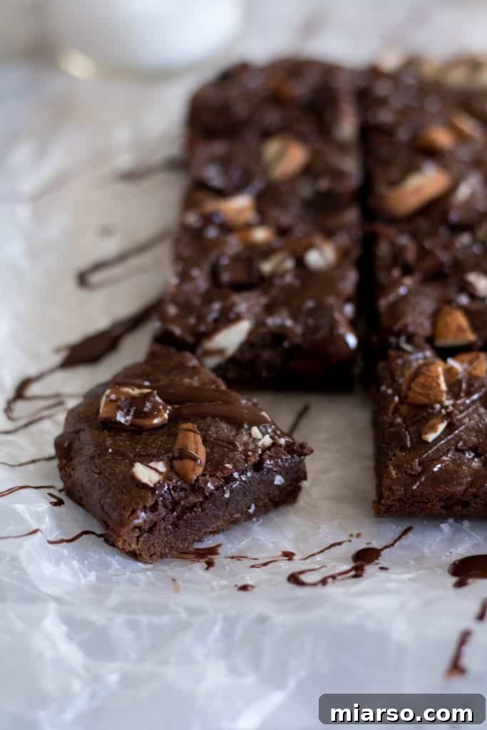 Delicious fudgy vegan double chocolate brownies, perfectly sliced into squares on parchment paper, topped with chopped almonds and a delicate chocolate drizzle, with one piece enticingly pulled away.
