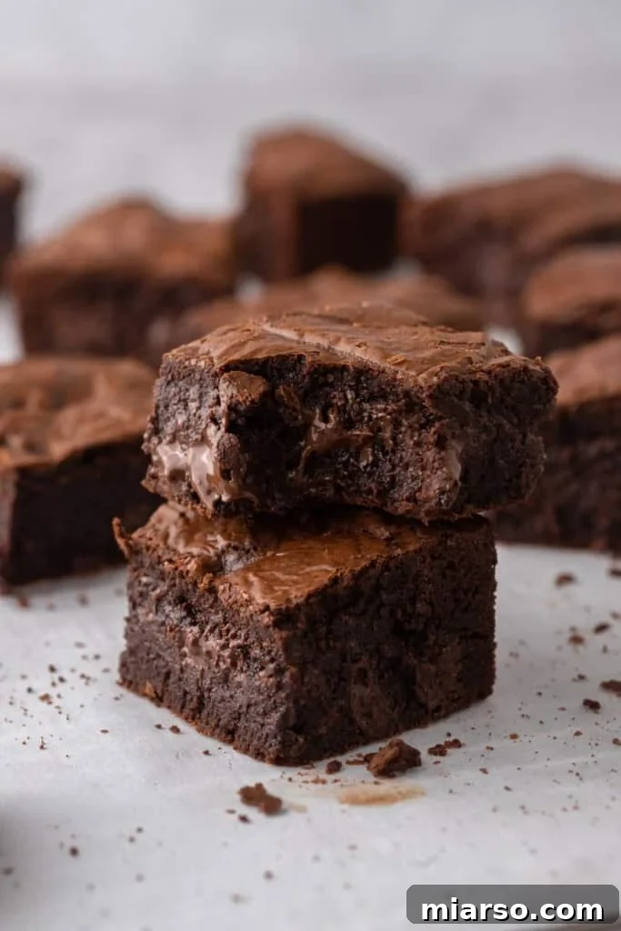 Two irresistibly fudgy brown butter brownies stacked on parchment paper, showcasing visible melted chocolate chunks within, with more decadent brownies blurred in the background.