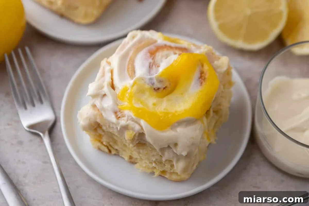 Lemon roll with lemon curd and cream cheese frosting on a white plate with lemons and a glass of frosting nearby