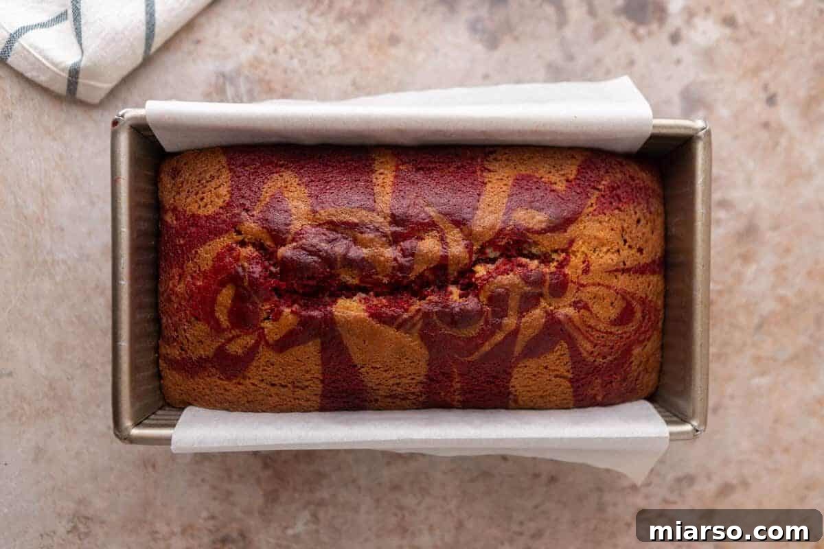 A freshly baked Red Velvet Marble Loaf Cake, still warm in its parchment-lined loaf pan, showcasing beautiful red and golden swirls on its perfectly domed top.