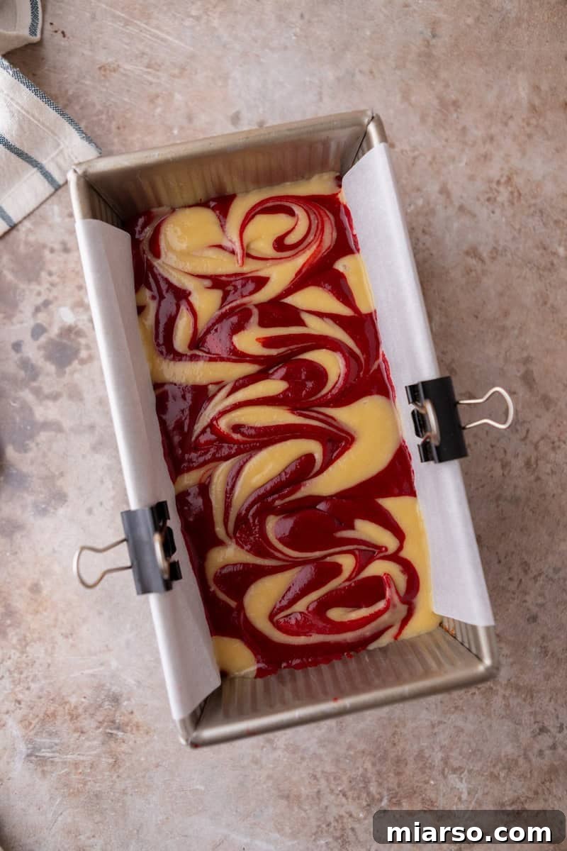 An overhead view of the marbled cake batter in a loaf pan after a single, gentle swirl with a sharp knife, revealing clear, contrasting patterns of red and vanilla before baking.
