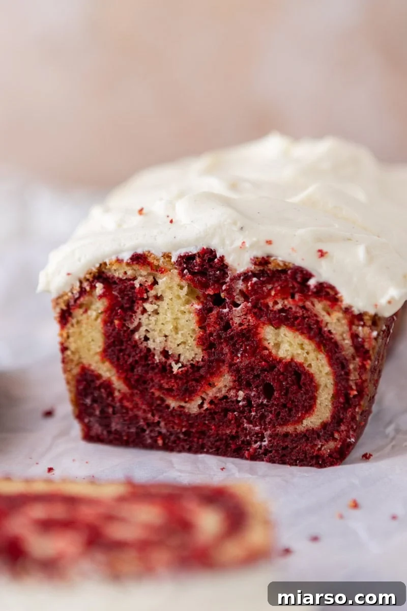 A straight, eye-level view of a perfectly frosted Red Velvet Marble Loaf Cake, adorned with a thick, artfully swirled layer of cream cheese frosting, clearly revealing the distinct marbled layers of red and vanilla cake beneath.