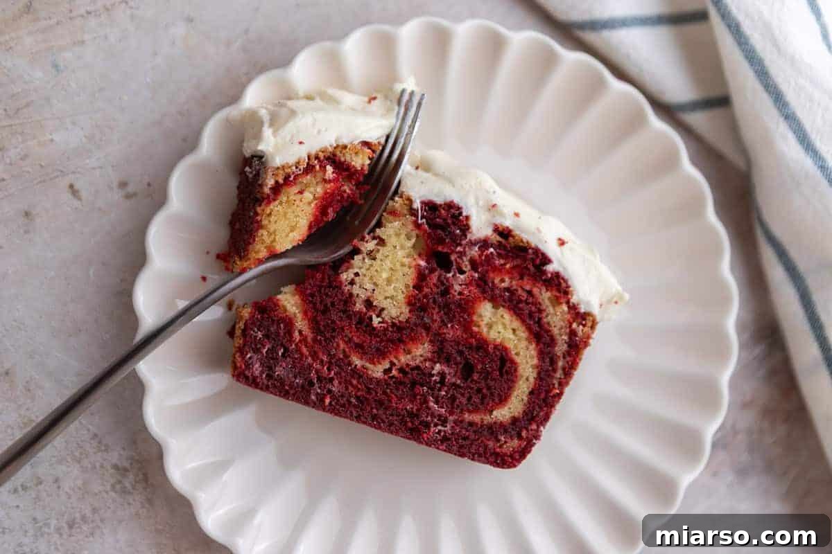 A thick, inviting slice of NY-style Red Velvet Marble Loaf Cake sits on a white plate, with a fork gently breaking into its tender, moist crumb, highlighting its dense texture and beautiful swirls.