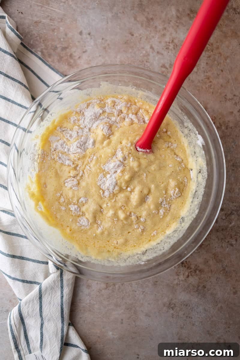 Ultimate Moist Marble Layer Cake with Rich Chocolate Cream Cheese Frosting 7 Wet ingredients being folded into dry ingredients with a red spatula in a glass bowl