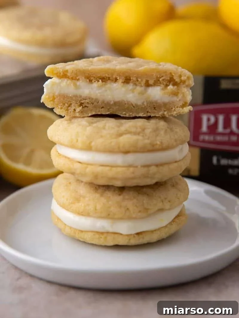 Stack of lemon sandwich cookies with cream cheese filling, top cookie split to show the inside, with lemons and Plugrà butter in the background.