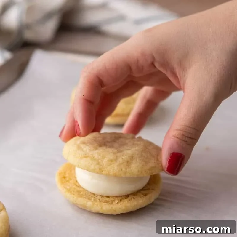 A hand placing the top cookie onto a cream cheese frosted lemon sandwich cookie to assemble it.