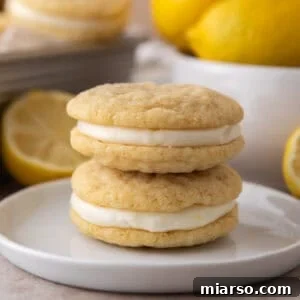 Two lemon sandwich cookies stacked on a small white plate with fresh lemons in the background.