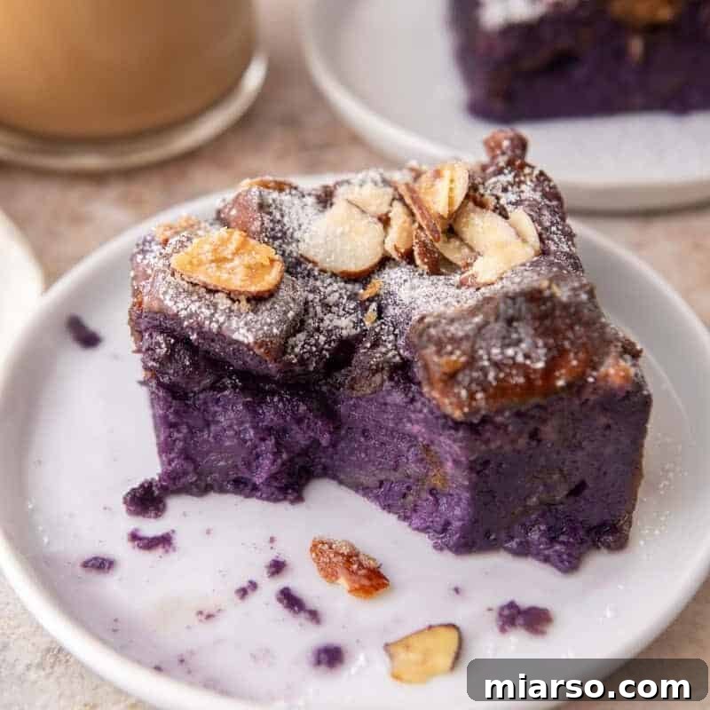 Ube French toast bake slice on a small white plate dusted with powdered sugar and toasted almonds with coffee in the background