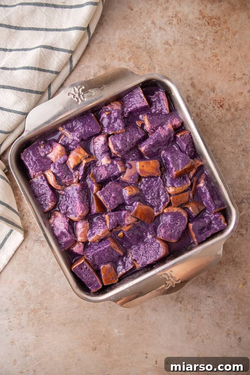 Cubed Hawaiian rolls soaking in purple coconut milk custard in a square metal baking dish