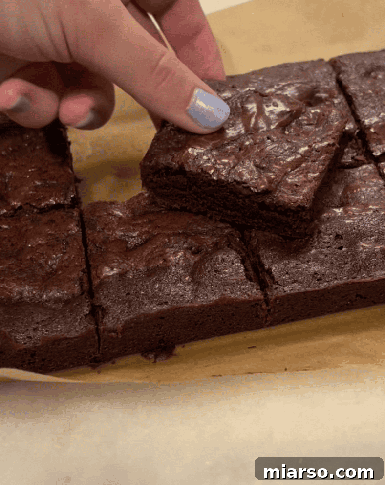 Hand reaching for a fudgy chocolate brownie square cut on parchment paper for Loaf BakeClub