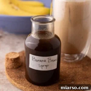 Banana bread syrup in a glass bottle with a handwritten label on a cork trivet next to an iced banana bread latte