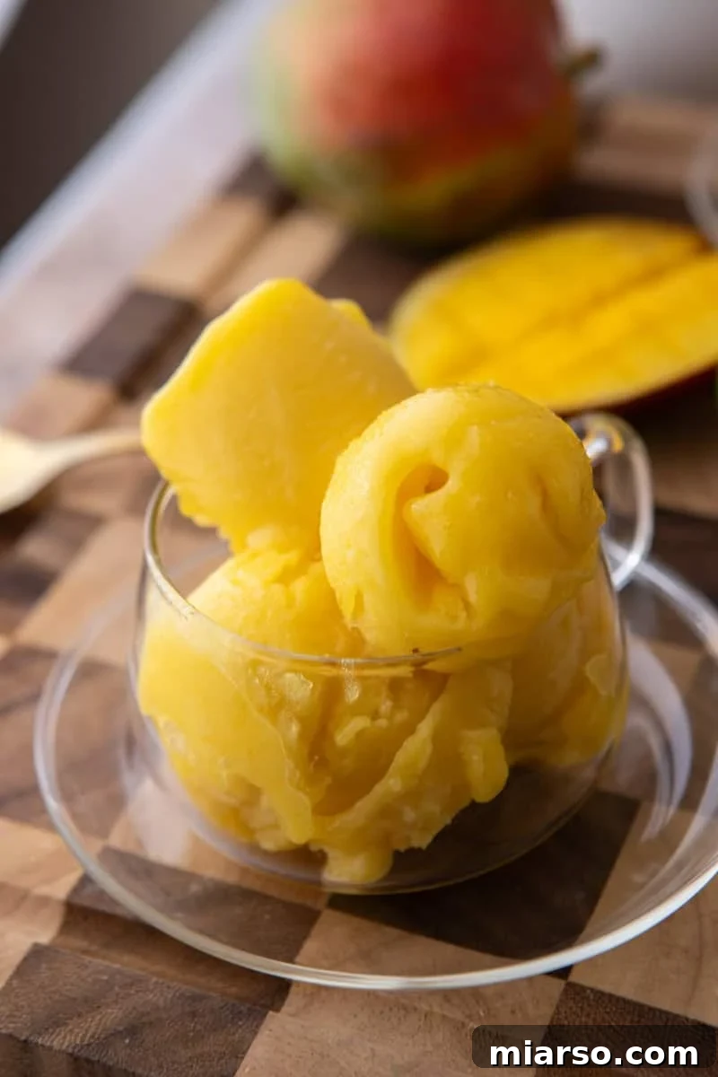  Scoops of velvety mango sorbet in a clear glass cup with fresh mango in the background