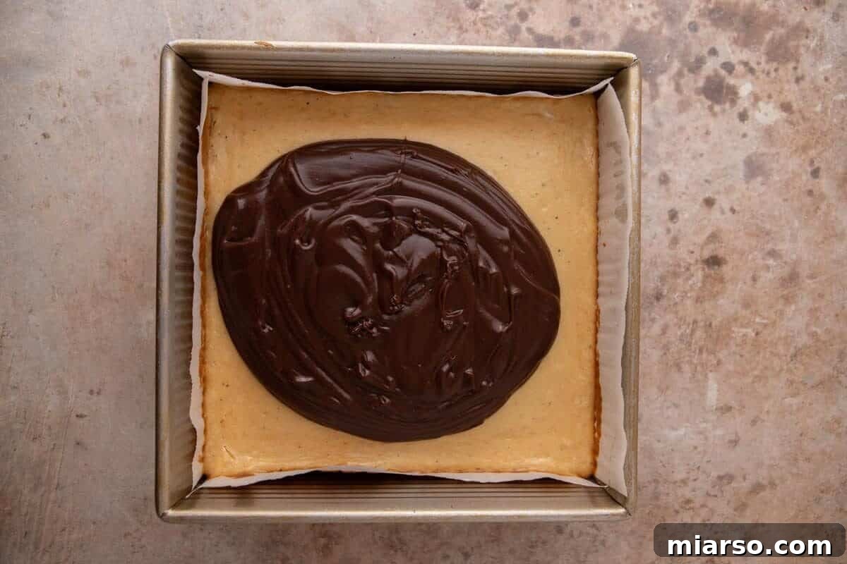 Chocolate ganache being spread over the baked marshmallow cheesecake layer for s'mores cheesecake bars
