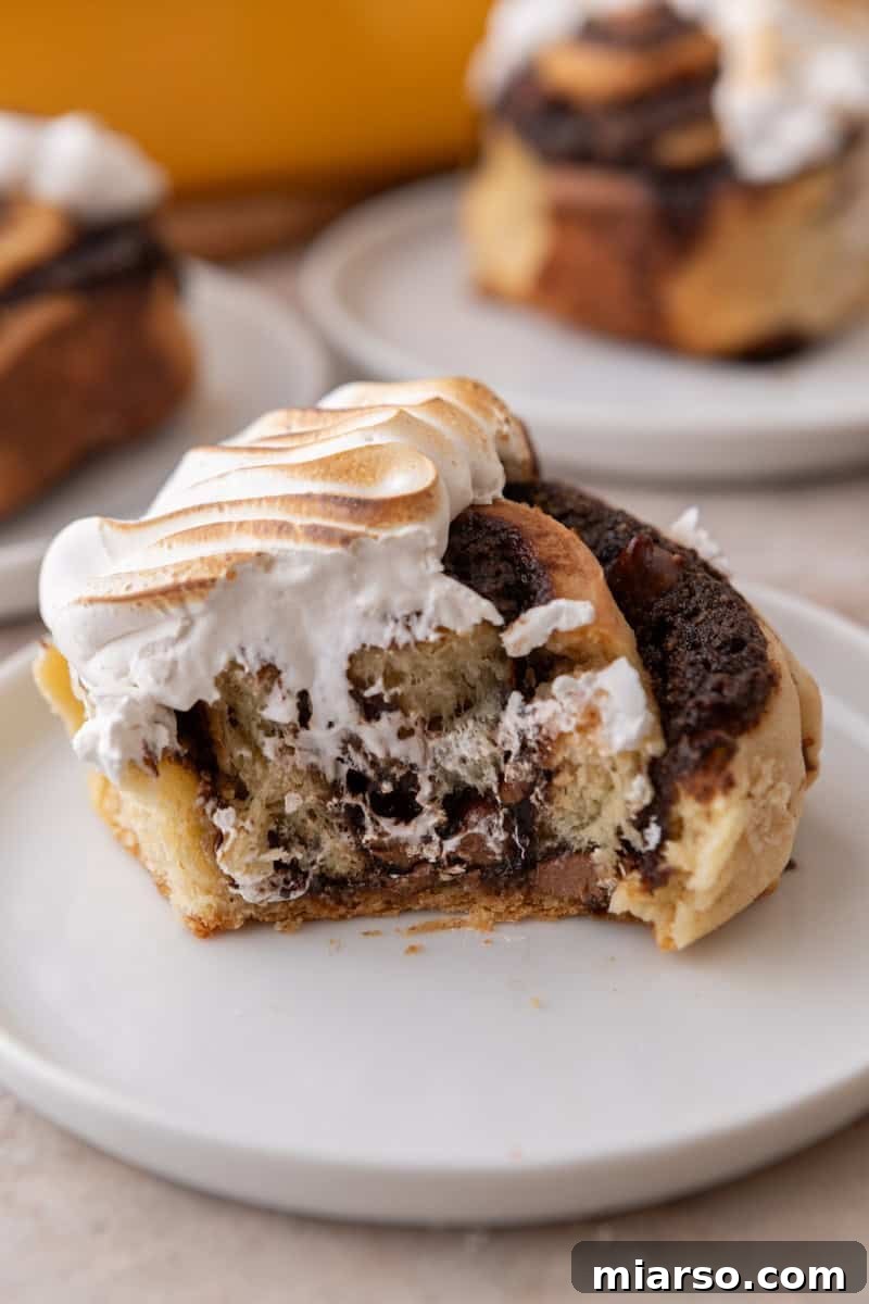S'mores cinnamon roll topped with toasted meringue on a white plate with a bite taken out