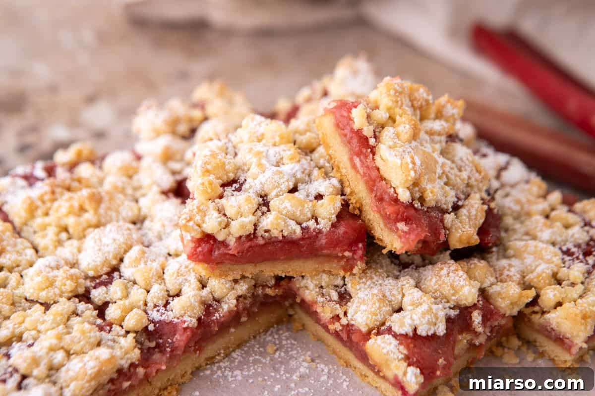 Tangy Rhubarb Crumble Bars with Oat Streusel 2 Rhubarb crumble bars stacked on a pink surface dusted with powdered sugar, with fresh rhubarb stalks in the background
