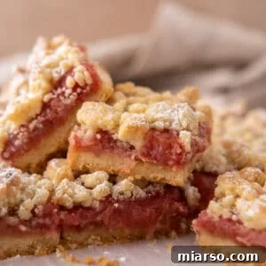 Stack of rhubarb crumble bars on parchment paper showing layers of golden shortbread base, pink jammy rhubarb filling, and crumble topping
