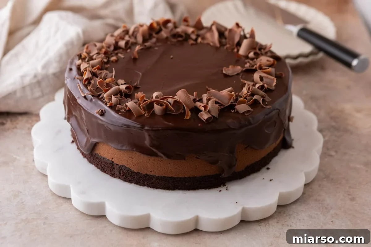 Decadent Chocolate Mousse Cake Recipe 2 Chocolate mousse cake on a white scalloped marble cake stand, topped with chocolate ganache and chocolate curls, with a cake server in the background