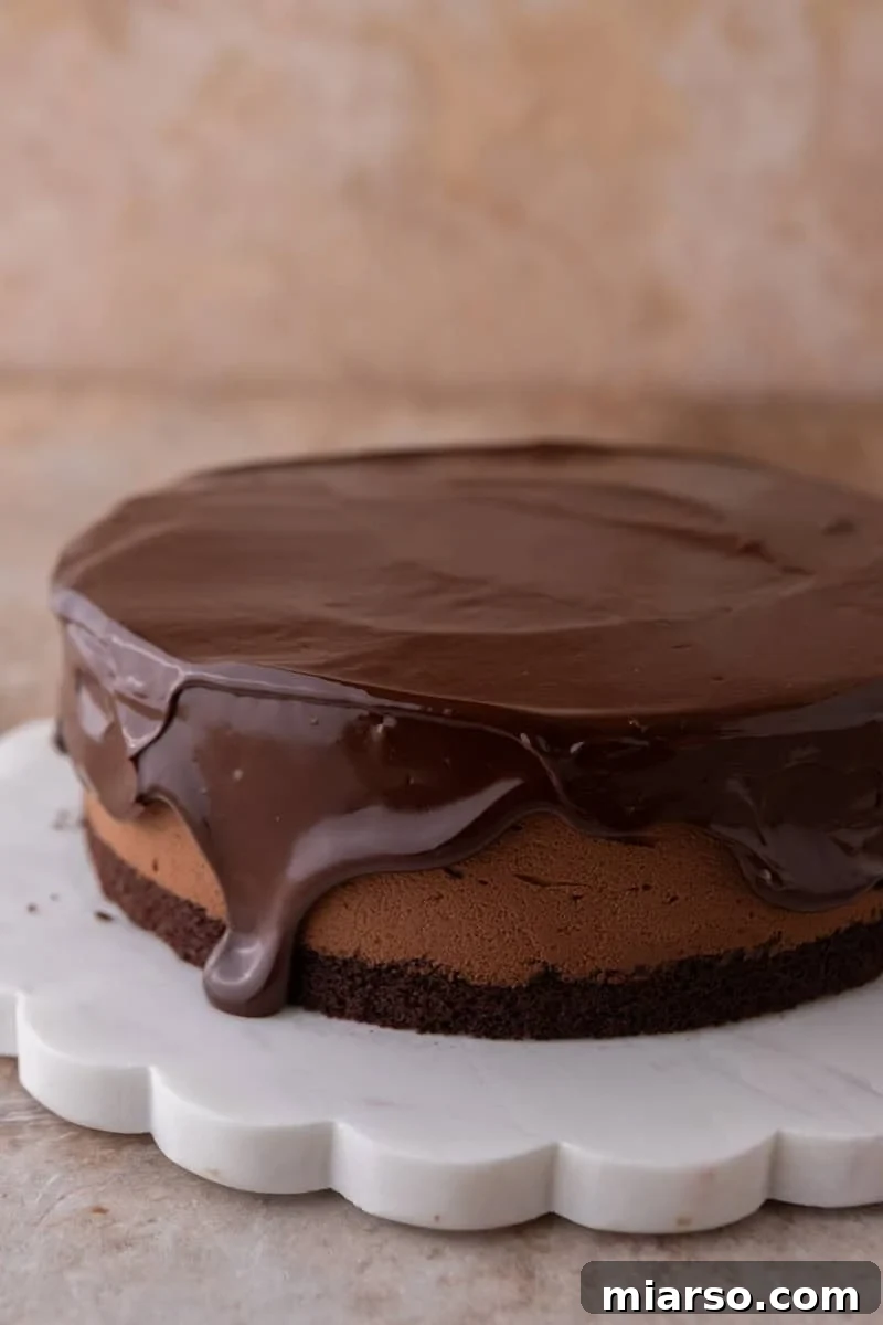 Decadent Chocolate Mousse Cake Recipe 14 Chocolate ganache being poured over the chocolate mousse cake, dripping down the sides, on a white scalloped marble cake stand