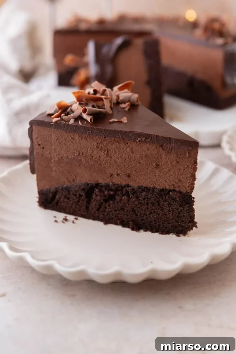Decadent Chocolate Mousse Cake Recipe 17 A slice of chocolate mousse cake on a white scalloped plate showing three distinct layers of chocolate sponge cake, chocolate mousse, and chocolate ganache topped with chocolate curls