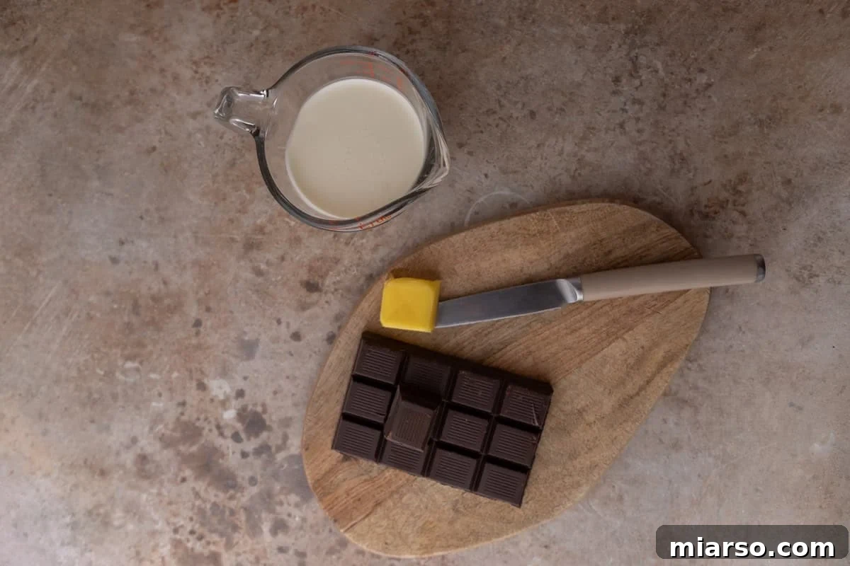 Decadent Chocolate Mousse Cake Recipe 5 Ingredients for the chocolate ganache including heavy cream in a glass measuring cup, a dark chocolate bar, and butter on a wooden cutting board